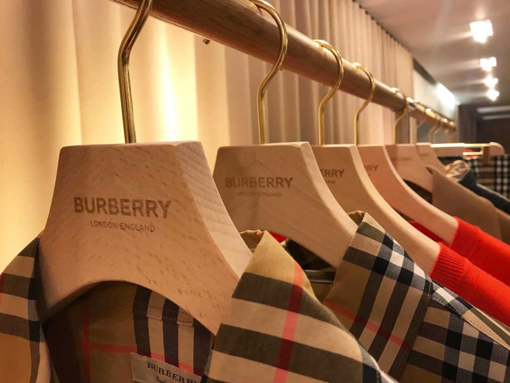 HOL Group Wooden hangers with "BURBERRY LONDON, ENGLAND" engraved on them hold a variety of Burberry clothing items, including the brand’s signature plaid patterns. Garments are hung in a neat row, with warm lighting in the background creating a luxurious ambiance.