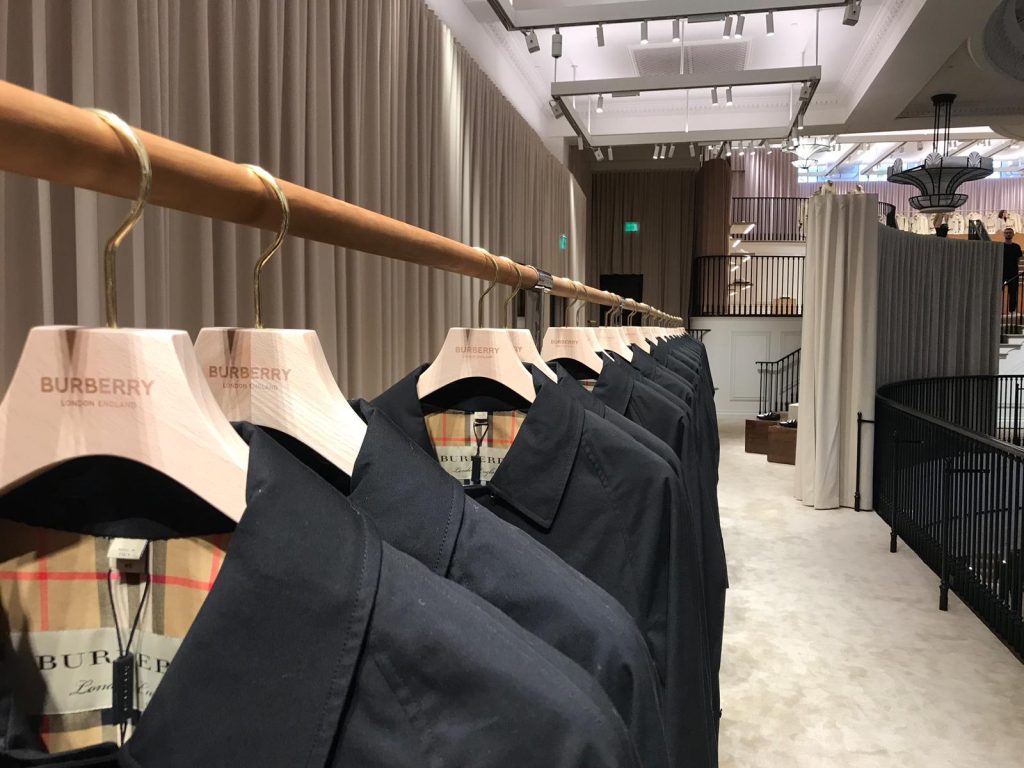 HOL Group A row of Burberry trench coats hangs on wooden hangers with Burberry branding. The coats are neatly displayed on a wooden rack in a well-lit, upscale retail store with beige curtains in the background and a modern, elegant interior.