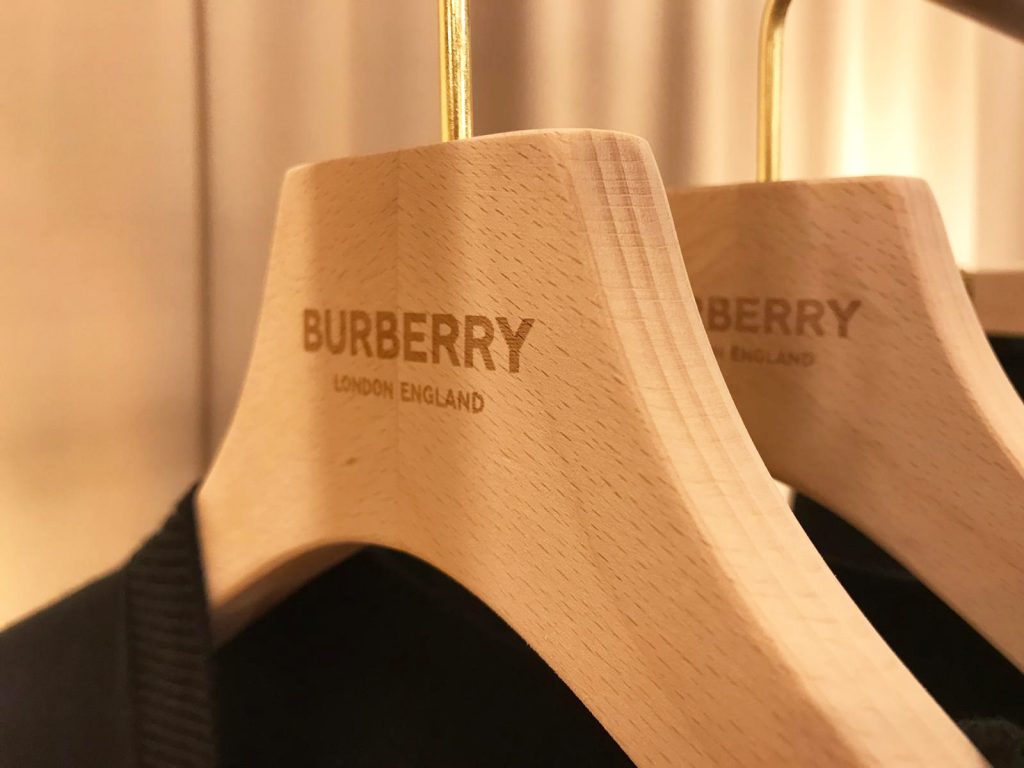HOL Group Close-up of wooden hangers branded with the "Burberry London England" logo in gold text. The hangers are holding dark-colored clothing and are set against a blurred background with warm lighting.
