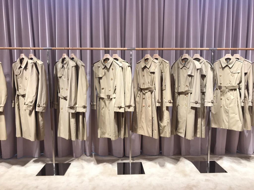 HOL Group A row of beige trench coats hangs on metal racks in front of a purple curtain backdrop. The coats, varying slightly in style, are neatly displayed in a well-lit retail setting.