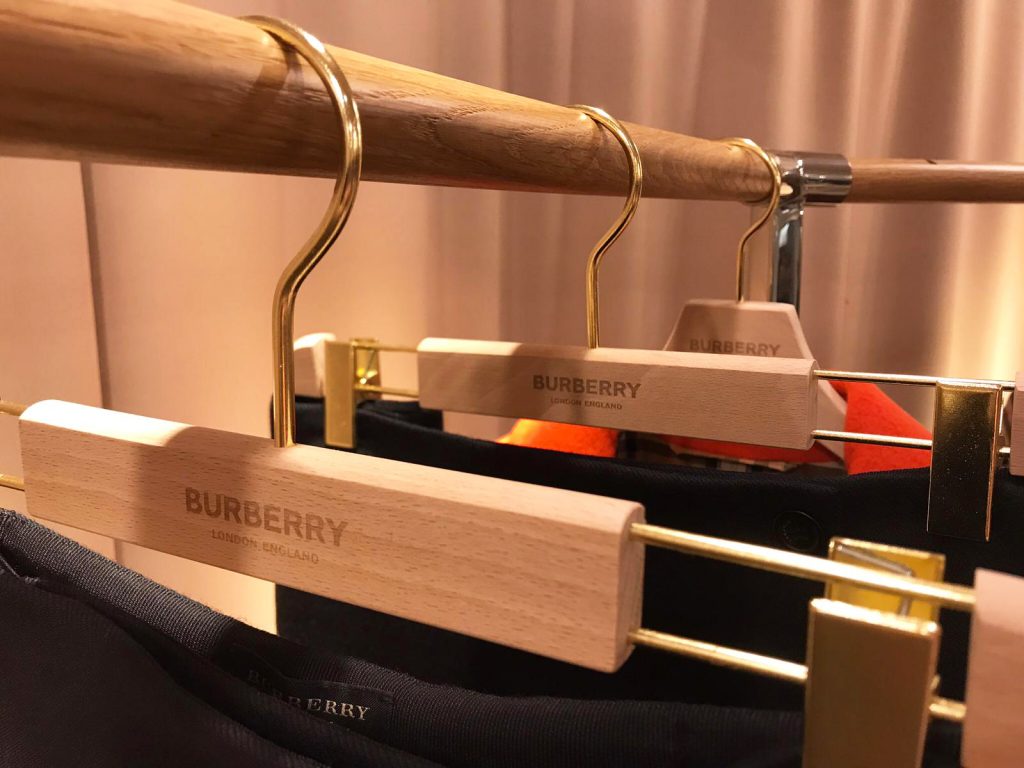 HOL Group Wooden clothes hangers branded with "Burberry" are hanging from a wooden rail. The hangers are holding dark-colored garments, and the setting appears to be a clothing store with warm lighting.