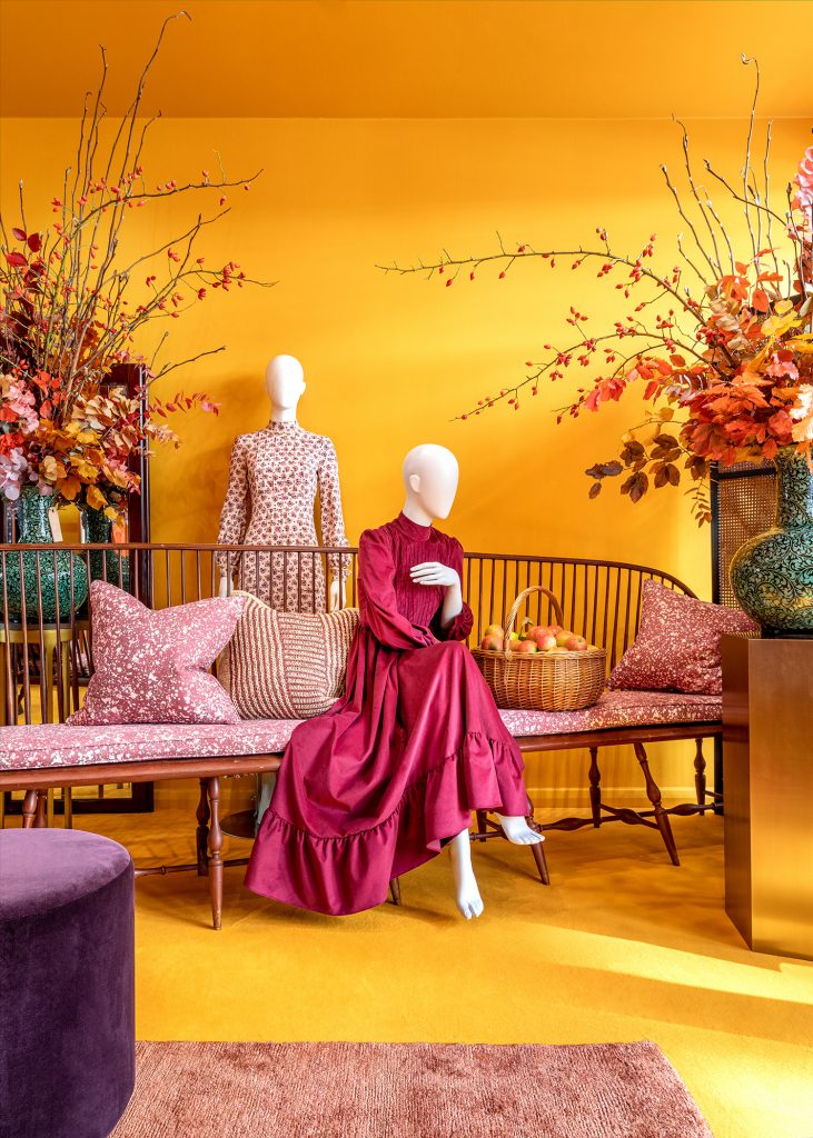 HOL Group A vibrant room features two mannequins from the Dress Edit collection, one in a deep red vintage-style dress seated, the other in a patterned daily dress standing. They are surrounded by plush cushions on a wooden bench and large vases of colorful autumn branches against a bright yellow backdrop.