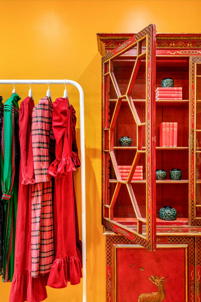 HOL Group A brightly colored room with an orange wall features a white garment rack holding four vibrant dresses in shades of red and green, perfect for your Daily Dress Edit. Beside the rack is an ornate red cabinet with geometric glass doors, displaying red books and decorative items.