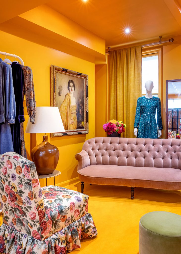 HOL Group A stylish room with bright yellow walls features a tufted blush sofa, a floral armchair, a brown lamp, and a mannequin dressed in this season's Daily Dress. A large painting of a woman in a yellow dress adorns the wall, serving as an elegant fashion edit, and a rack of clothes is visible on the left side.