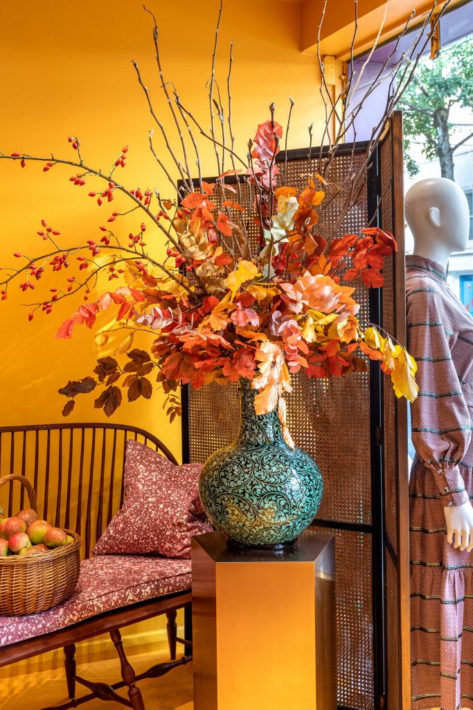 HOL Group A tall, intricately designed blue vase filled with colorful autumn leaves and branches sits on a pedestal in a vibrant room with orange walls. To the side, a basket of apples is placed on a bench with floral cushions, and a mannequin showcasing the Daily Dress Edit in plaid stands nearby.