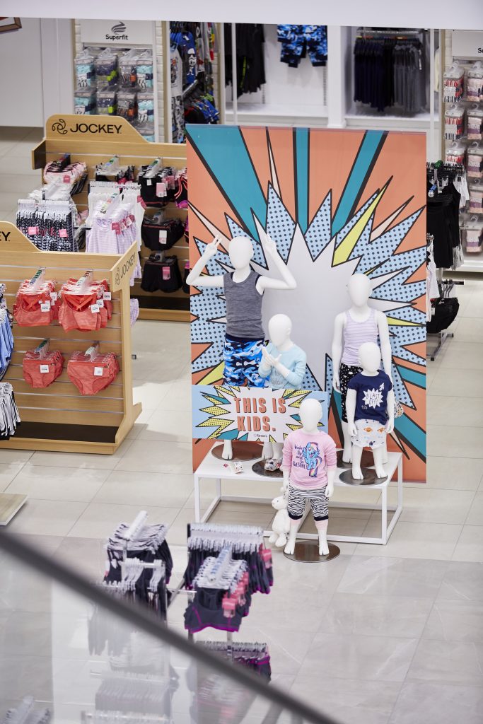 HOL Group A store display featuring white mannequins dressed in kids' clothing in front of a colorful, comic-style backdrop with the text “This is Kids.” Surrounding shelves showcase various items, from kids' underwear and socks to charming farmers-themed accessories.