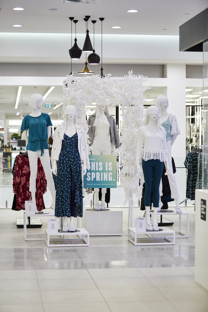 HOL Group A store display showcases mannequins dressed in spring fashion, including dresses, blouses, pants, and jackets in light colors. A sign reads "This is Spring," evoking the freshness of farming fields. The white decor and bright lighting highlight the modern, farm-inspired style.