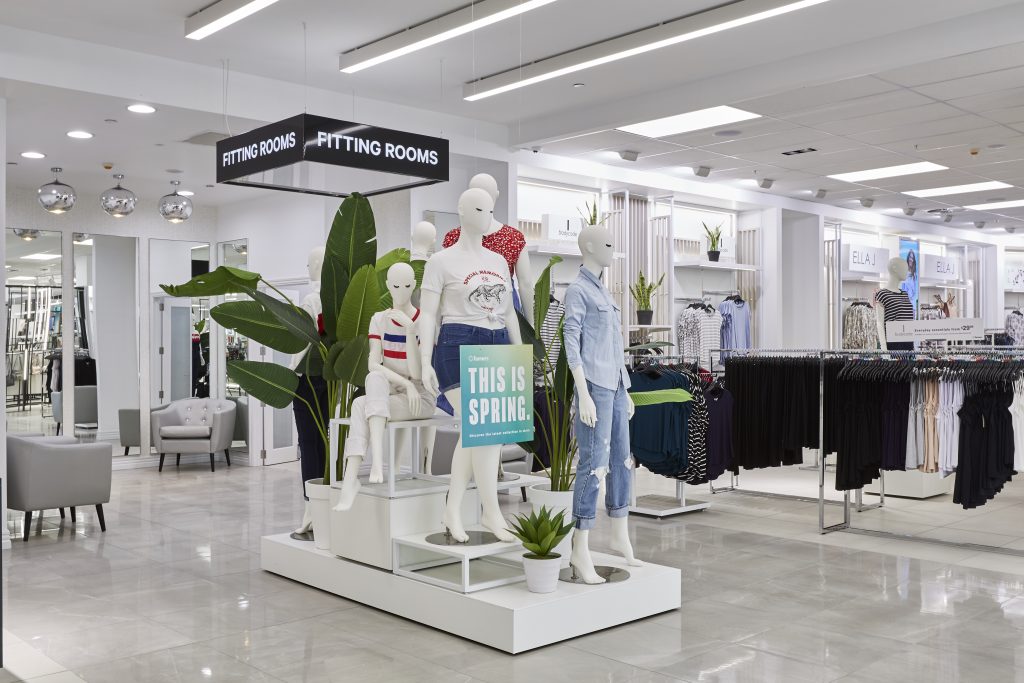 HOL Group A well-lit clothing store interior showcasing a display of mannequins dressed in spring outfits, reminiscent of farmers' markets. The mannequins hold a sign that reads "This is Spring." The background features racks of neatly arranged clothes, fitting rooms, and indoor plants.