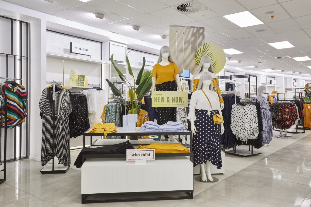 HOL Group A modern retail store features mannequins dressed in bold, stylish outfits, highlighted by a yellow top and striped skirt. Clothing racks display various colorful garments, and signs read "NEW & NOW" and "wild child." The store's bright, welcoming atmosphere appeals to urban go-getters and fashion-forward farmers alike.