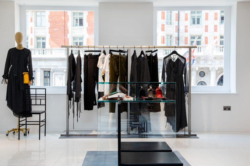 HOL Group A modern boutique, reminiscent of a luxury department store like Harvey Nichols, showcases a minimalist interior with two large windows. Various clothing items such as black dresses and pants are displayed on racks. A glass display case in the center holds accessories like bags and shoes. A mannequin stands on the left side.