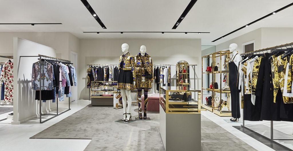HOL Group A luxurious clothing store interior reminiscent of Harvey Nichols, with mannequins dressed in patterned outfits on display. Racks of designer clothes line the walls, and shelves hold accessories and handbags. The decor is modern, blending white, gold, and neutral tones in true luxury department store style.
