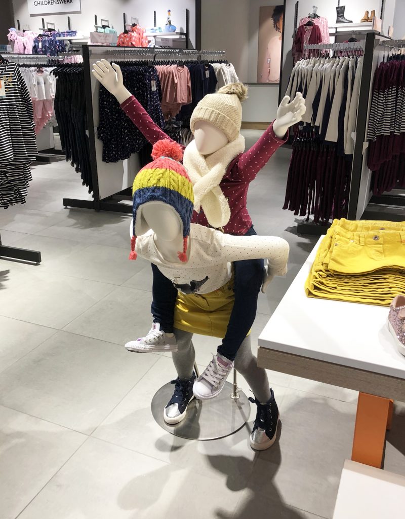 HOL Group Two child mannequins in a John Lewis clothing store, with one mannequin sitting on the other's shoulders. Both mannequins are dressed in winter outfits, including hats and scarves. Shelves and racks filled with children's clothing can be seen in the background.