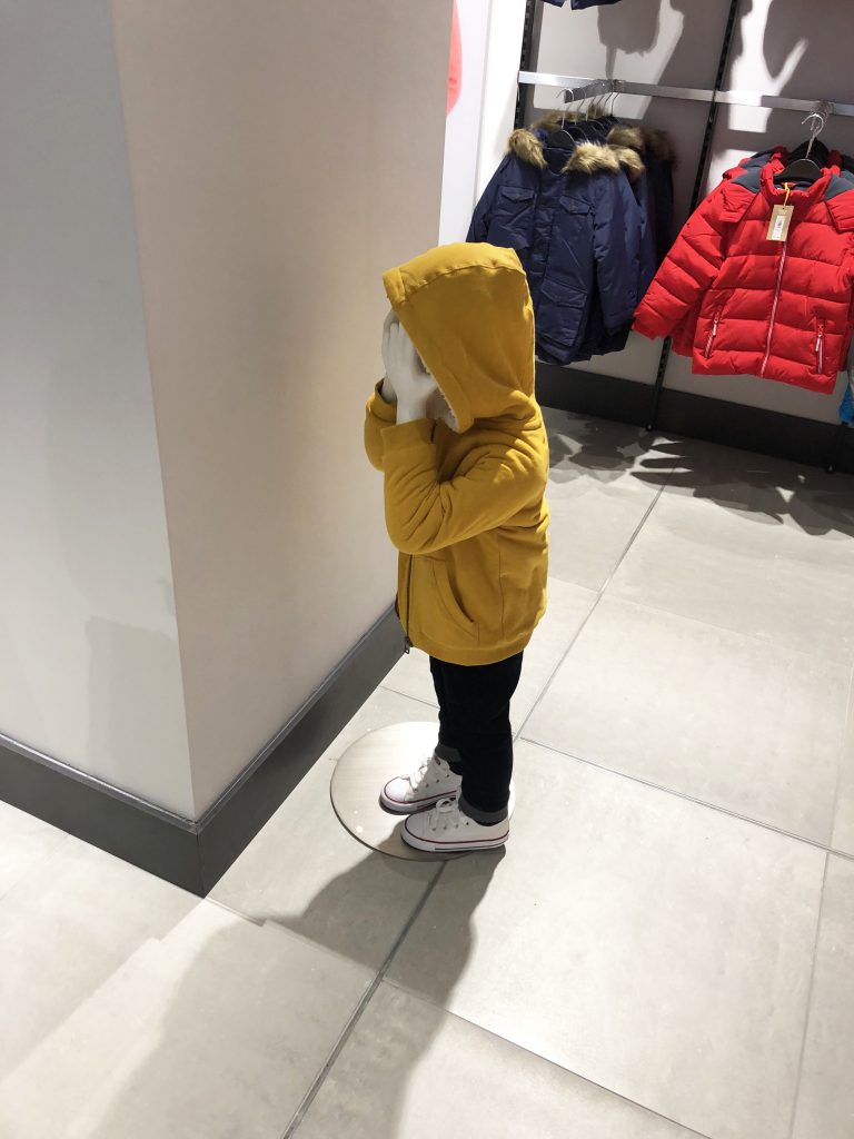 HOL Group A child mannequin dressed in a yellow hooded jacket, dark pants, and white sneakers stands facing a wall with hands over its face in the John Lewis clothing store. Coats are hanging on racks in the background.