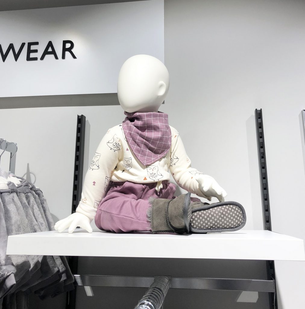 HOL Group A faceless mannequin dressed in a white top with printed graphics, mauve pants, a matching mauve bib, and gray boots is posed sitting cross-legged on a white display shelf in a John Lewis store. The background features a partial clothing rack and a sign that reads "WEAR.