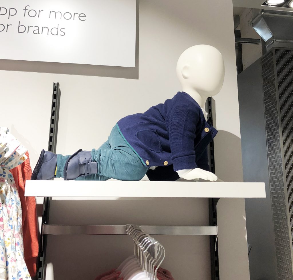 HOL Group A faceless mannequin in a John Lewis children's clothing store is posed on all fours on a display shelf. The mannequin is dressed in a navy blue sweater, teal pants, and matching dark blue boots. Hanging clothes and a signboard are visible in the background.
