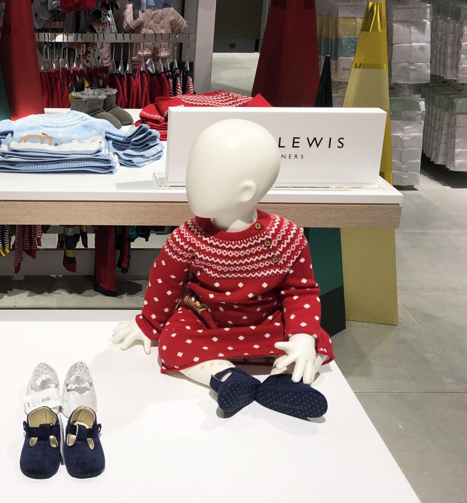 HOL Group A headless mannequin dressed in a red and white patterned sweater and red pants sits on a display table in the John Lewis children’s clothing store. In front of the mannequin, there are pairs of children's shoes. Folded clothes are stacked neatly in the background.