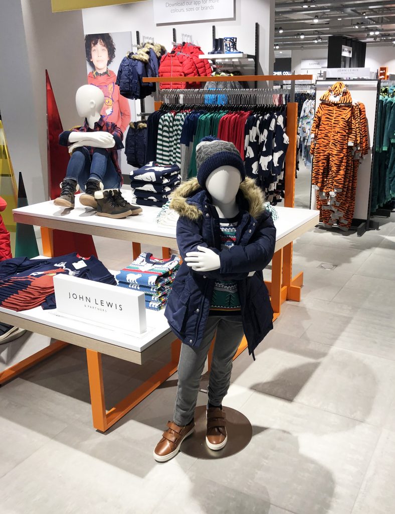 HOL Group A John Lewis retail store display features two child mannequins dressed in winter clothing. One sits on a table wearing a red jacket, jeans, and boots. The other stands wearing a navy coat, gray pants, striped sweater, beanie, and brown boots. Racks of clothes are in the background.