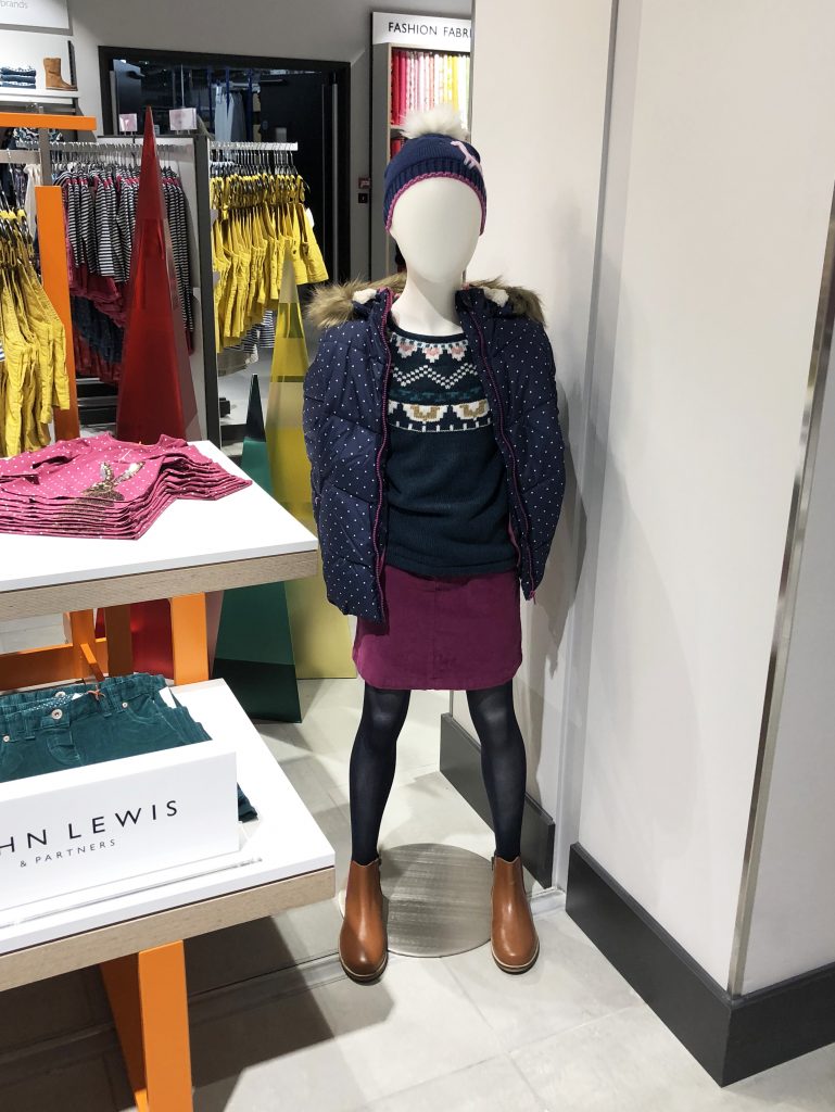 HOL Group A John Lewis store mannequin is dressed in a navy polka-dot puffer jacket, a patterned sweater, a maroon skirt, black tights, brown boots, and a colorful knit hat. The display stands near a stack of folded clothes and vibrant yellow pants in the background.