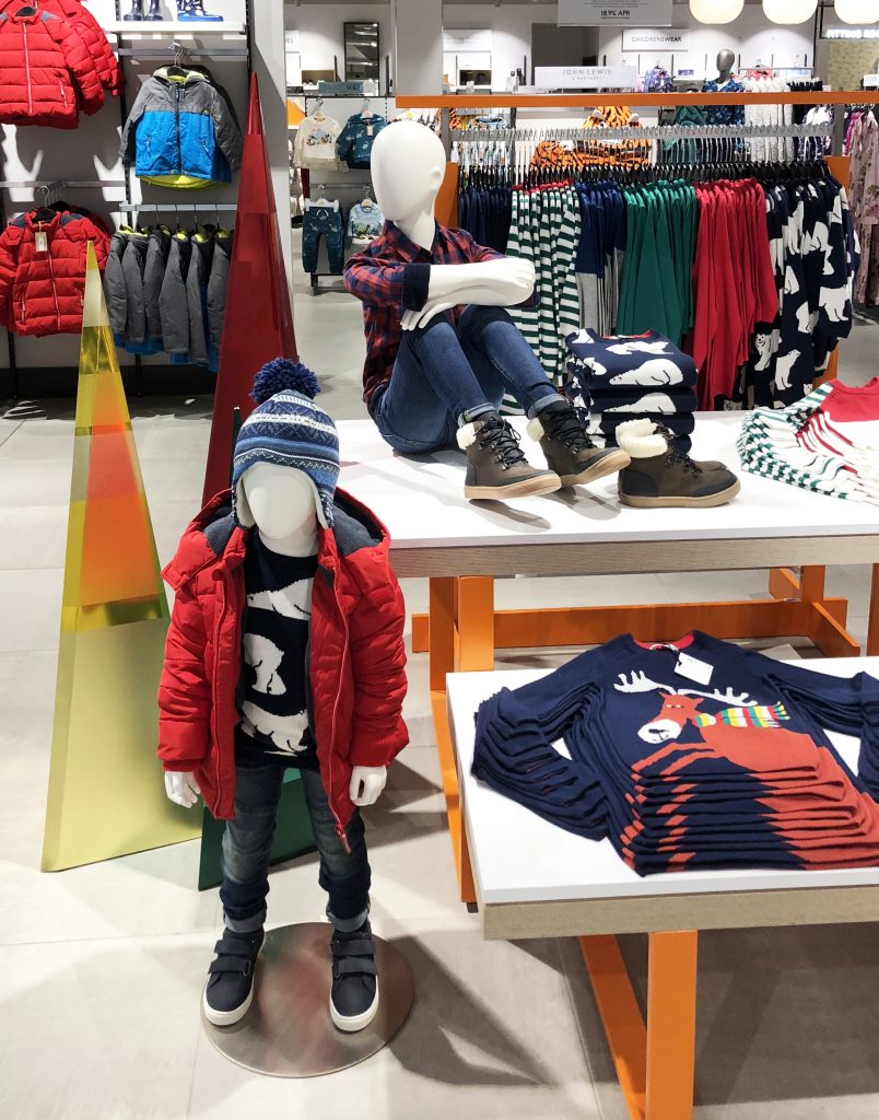 HOL Group A display in a John Lewis clothing store features child-sized mannequins in winter outfits. One mannequin sits atop a table wearing a flannel shirt, jeans, and boots, while another stands below in a red jacket and hat. Folded clothes are neatly arranged around them.