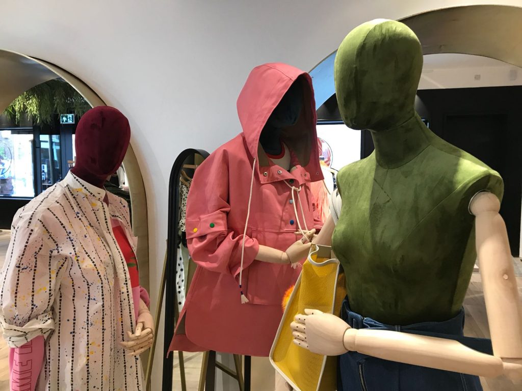 HOL Group Three mannequins dressed in colorful, modern outfits are displayed in a stylish, well-lit emporium. The foreground mannequin wears a green top and holds a yellow bag. Another mannequin in a pink hooded jacket and one in a spotted outfit are visible in the background.