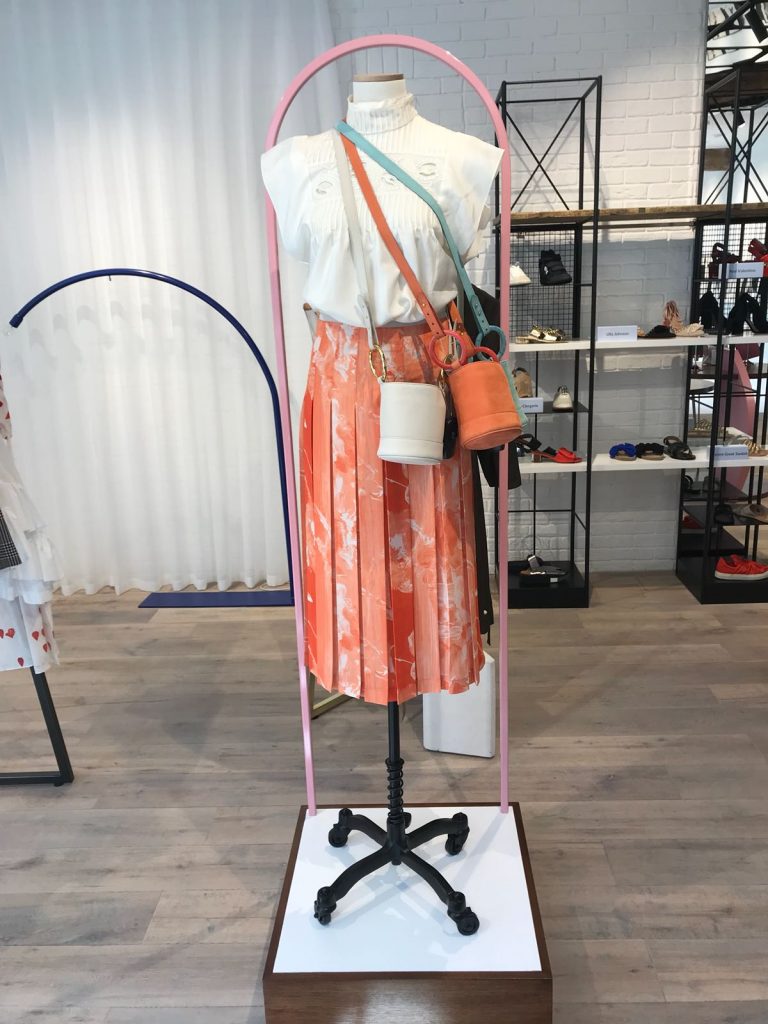 HOL Group A mannequin is dressed in a white sleeveless blouse and an orange pleated skirt with white patterns. It is adorned with a white cylindrical handbag and an orange bucket bag, along with a green crossbody bag. The vibrant background of the puzzle shop emporium showcases more accessories and puzzles.