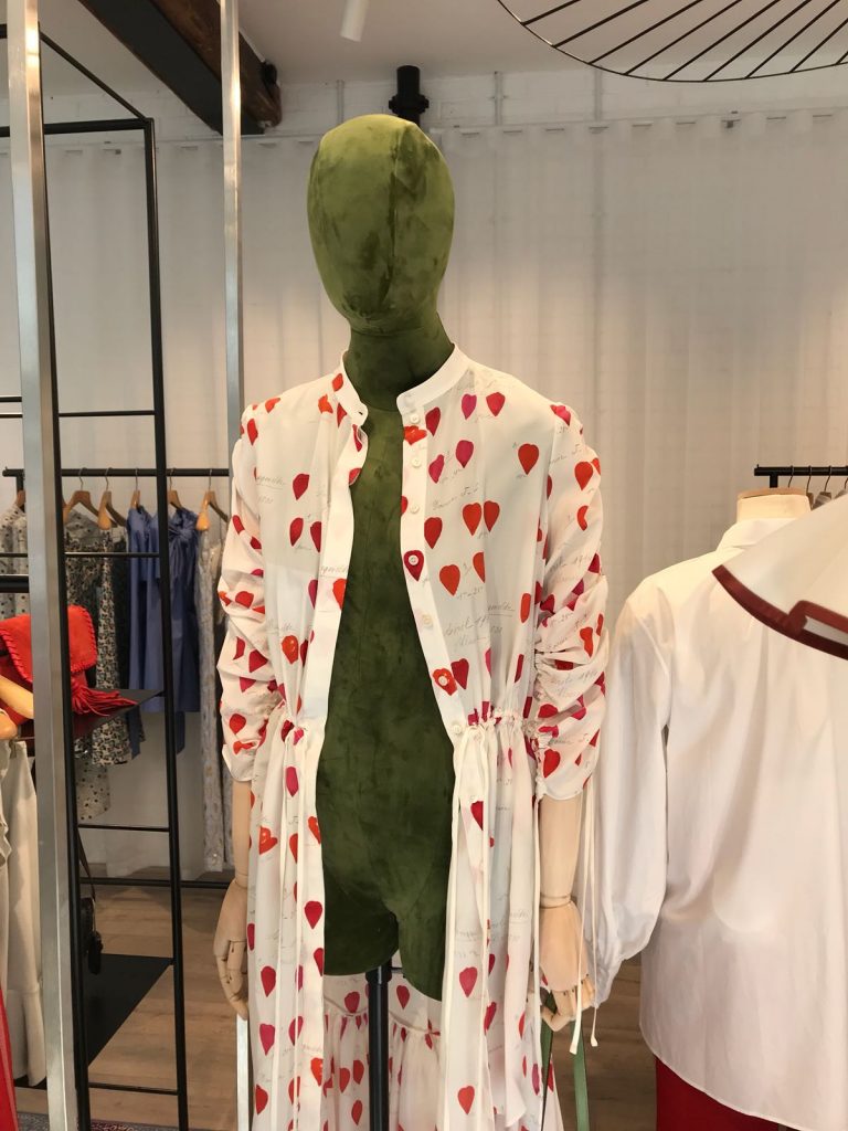 HOL Group A green mannequin is dressed in a white outfit adorned with red and orange heart patterns, featuring a long-sleeve top and matching pants. The mannequin stands prominently in the fashion emporium, with more clothing visible in the background.