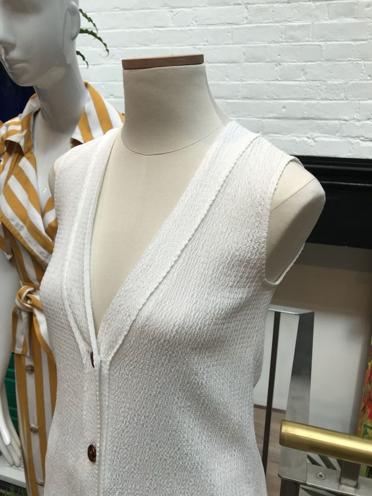 HOL Group A mannequin at the emporium displays a sleeveless white textured V-neck dress with button details. Another mannequin in the background is dressed in a yellow and white striped outfit with a tied waist. Both mannequins are in front of a white brick wall, creating a charming scene akin to pieces of an intricate jigsaw puzzle.
