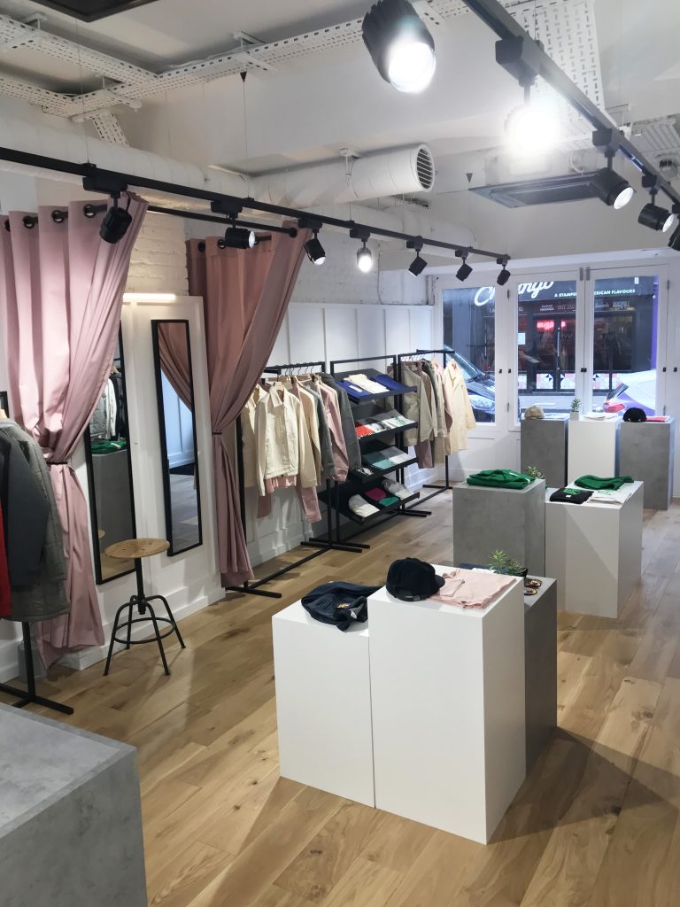 HOL Group A modern, minimalist clothing store with wooden floors and high ceilings. Clothing racks displaying various garments, including trendy workwear from MC Overalls, are against the walls. Two fitting rooms with pink curtains are on the left. Several display tables with folded clothes and accessories stand in the center.