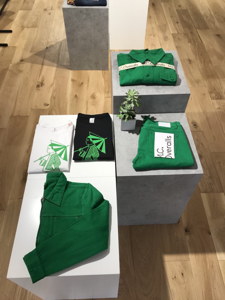 HOL Group A display of folded green and white clothing items on concrete pedestals showcases a set of green overalls, a white t-shirt with a green graphic, and a black t-shirt with a similar design. Evoking the essence of MC Overalls workwear, the setup is complemented by a small potted plant on the wooden floor.