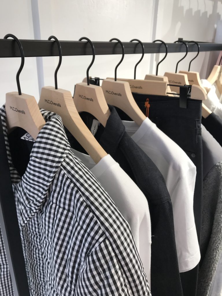 HOL Group A row of shirts on wooden hangers hangs on a clothing rack. The assortment includes an MC Overalls black and white checkered shirt, a black shirt, several white shirts, and other various colors. Each garment is neatly arranged, showcasing a minimalist and organized fashion display.