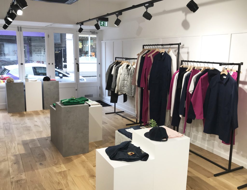 HOL Group A modern boutique store with light wooden floors and white walls. Clothing racks display various garments, including shirts, jackets, and MC Overalls in neutral and vibrant colors. Countertops hold folded clothes and hats, with large windows letting in natural light.
