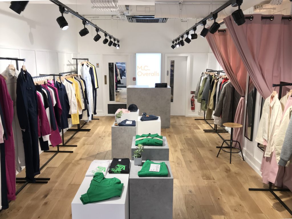 HOL Group A modern clothing store, featuring MC Overalls, has neatly arranged racks of colorful outfits on both sides, with tables in the center displaying folded garments. The store has wooden floors, white walls, and track lighting, creating a bright and inviting atmosphere. A curtain and fire extinguisher are visible.