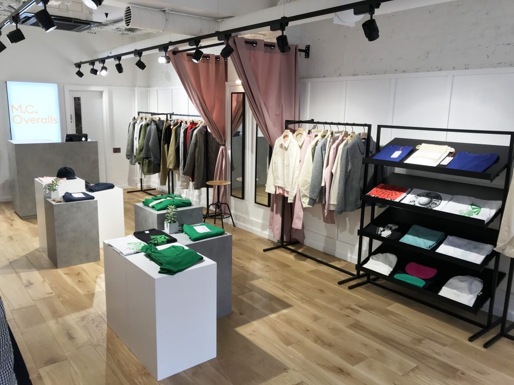 HOL Group A modern clothing store with wooden flooring and industrial-style lighting, featuring racks displaying various clothing items and shelves holding folded workwear. A pink curtain partially covers a fitting room. The counter at the back has a screen displaying "MC Overalls.