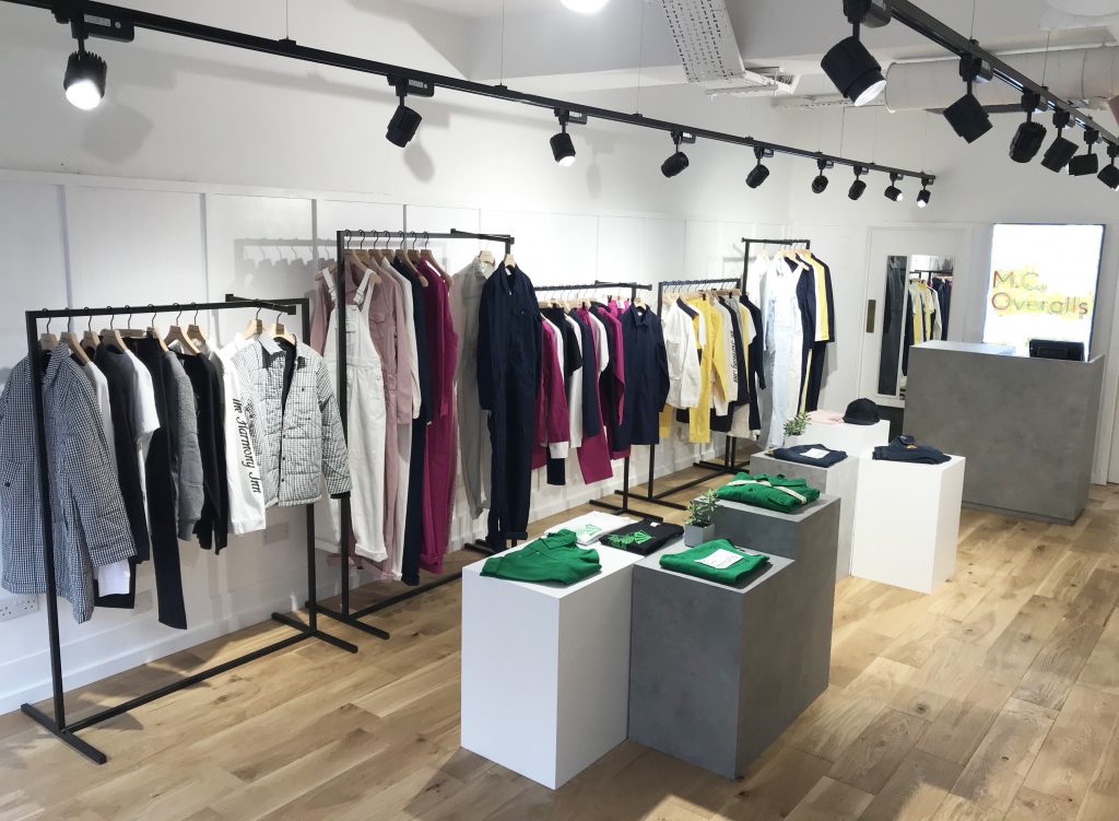HOL Group A modern clothing store featuring various garments on racks, including jackets, shirts, and pants in a range of colors such as black, white, pink, yellow, and green. Folded clothing items are displayed on minimalist grey tables. The floor is wooden. MC Overalls are prominently showcased.