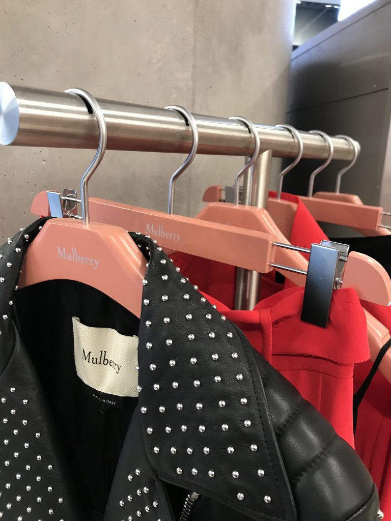 HOL Group A row of pink Mulberry hangers hold various garments on a metal clothing rack. The focus is on a black leather jacket adorned with silver studs. To the right, bright red clothing items, possibly dresses or jackets, are also visible.