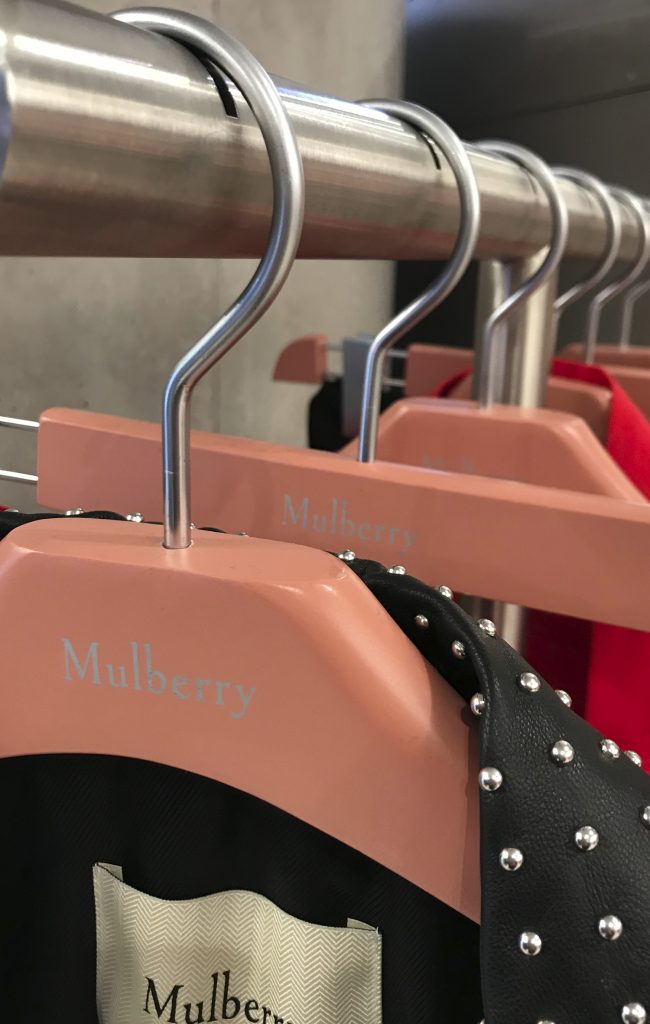 HOL Group Close-up image of a clothing rack displaying wooden hangers labeled "Mulberry." A studded black jacket and a vibrant red garment are partially visible hanging on the rack. The stylish setup suggests this is part of a chic Mulberry clothing store or showroom.