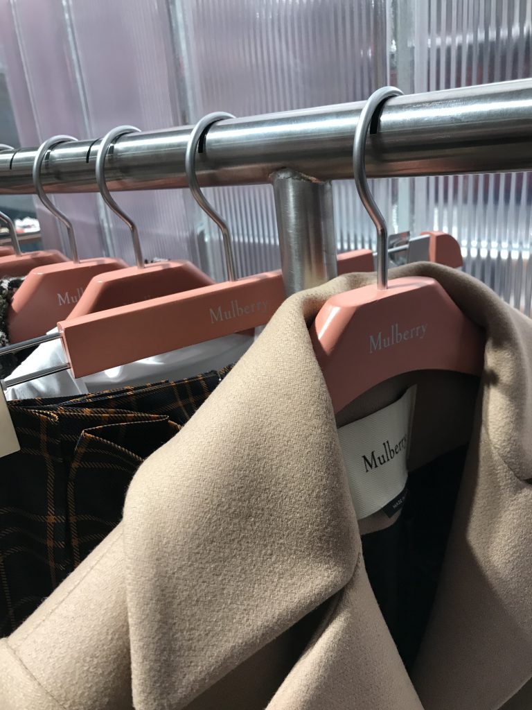 HOL Group A close-up view of a clothing rack showcasing several Mulberry garments on pink hangers. The beige coat in the foreground stands out, while more elegant clothing items can be seen in the background.