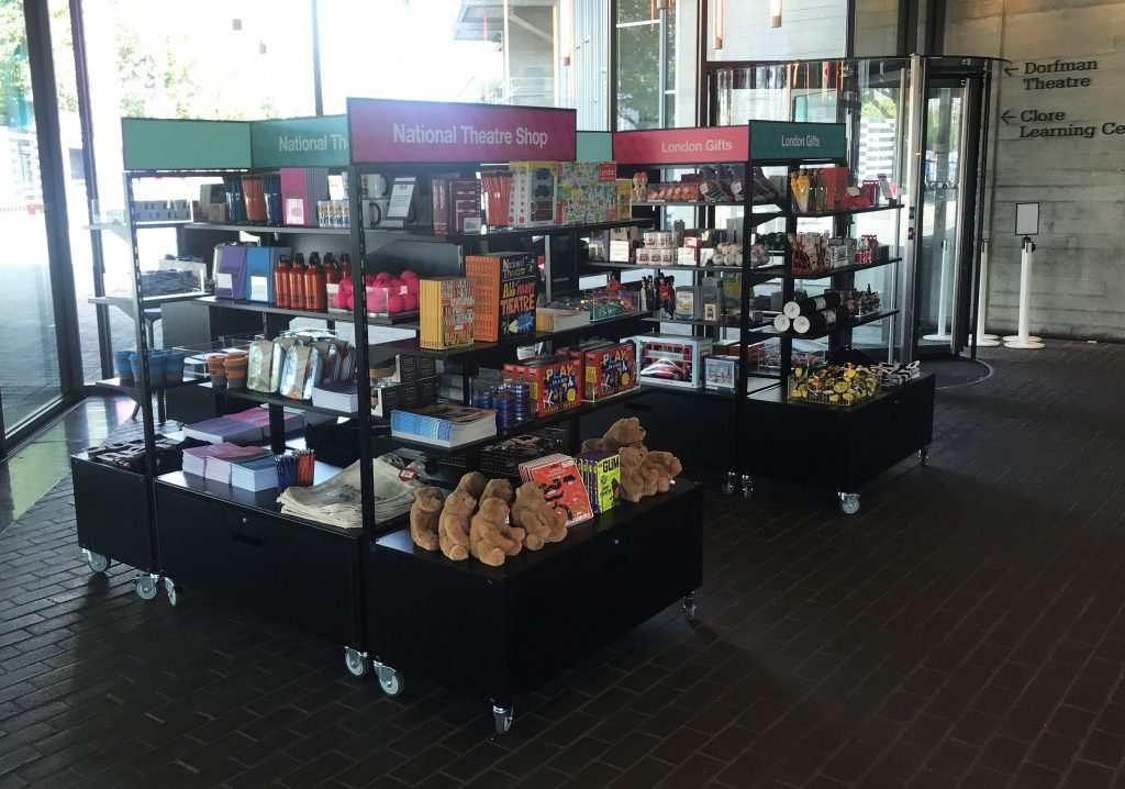 HOL Group A merchandise shop with two display stands selling various items such as souvenirs, toys, books, and gifts. The stands are labeled "National Theatre Shop" and "London Gifts." In the background, there is a circular glass entrance and signage for the Dorfman Theatre at the National Theatre complex.