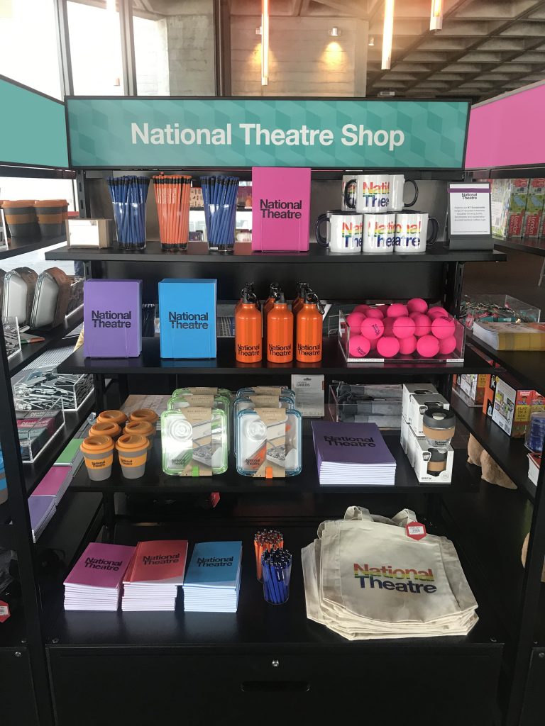 HOL Group A display in the National Theatre Shop features various merchandise, including branded books, notebooks, orange water bottles, pink stress balls, canvas tote bags, reusable coffee cups, pencils, and pens. The sign above proudly reads "National Theatre Shop".