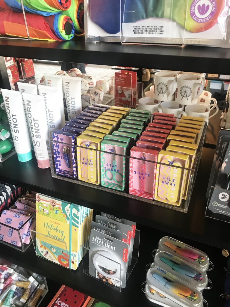 HOL Group A store display shelf filled with colorful items including tubes of hand cream, variously colored nail files, holiday notecards, highlighters, and other stationery and personal care products. The shelves are organized like the set design at the National Theatre, with each item neatly arranged.