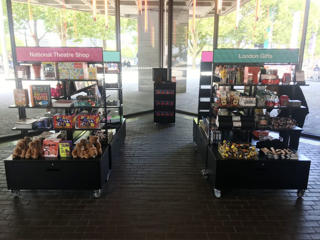 HOL Group An indoor shop features two main sections: "National Theatre Shop" on the left, offering an array of books, stuffed bears, and games relating to the National Theatre, and "London Gifts" on the right with various souvenirs like mugs, toys, and keychains. Large windows in the background reveal an outdoor area.