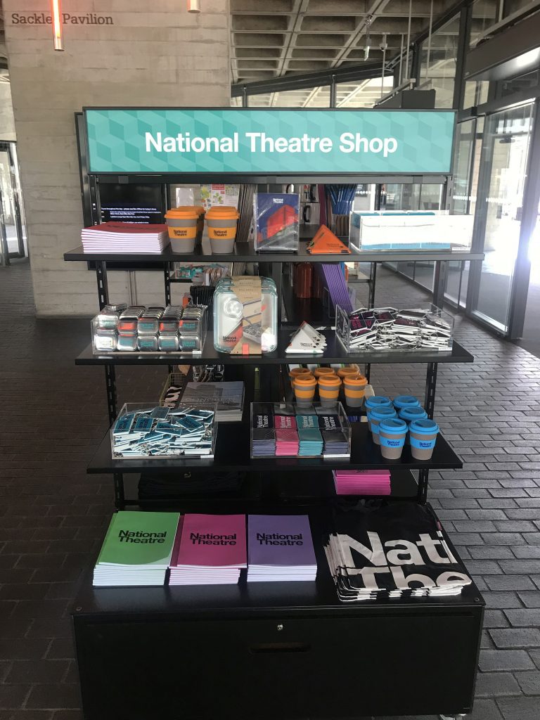 HOL Group A display rack at the National Theatre Shop holds various items, including mugs, books, and souvenirs. The top section features a sign labeled "National Theatre Shop." This charming store is located in a building with large glass windows and a concrete ceiling.