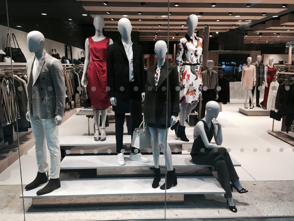 HOL Group A store window display features seven mannequins dressed in stylish Reiss clothing. Outfits include dresses, blazers, and casual wear in both neutral and vibrant colors. The background showcases neatly arranged clothing racks and shelves, creating a chic and modern atmosphere.
