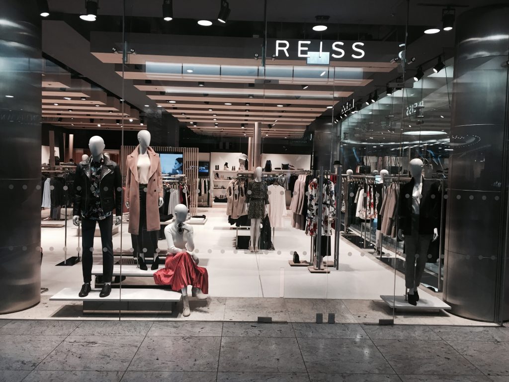 HOL Group A storefront display of the fashion brand Reiss shows mannequins dressed in stylish outfits, including jackets, coats, and skirts. The interior features neatly arranged racks of clothing and a polished, modern design with bright lighting.