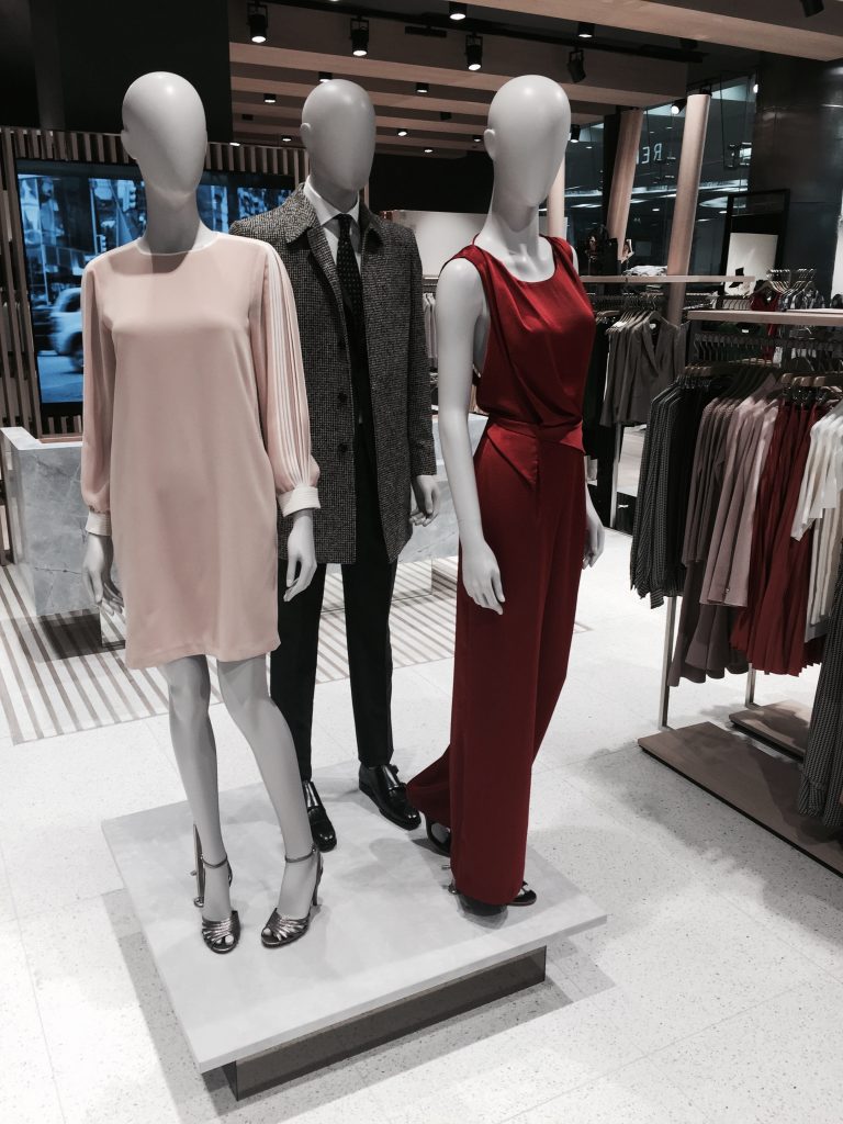 HOL Group Three mannequins are displayed in the Reiss clothing store. One mannequin wears a pink long-sleeve dress and heels, another sports a grey coat with a suit, and the third is dressed in a red sleeveless jumpsuit with heels. The background shows racks of clothes and the store interior.