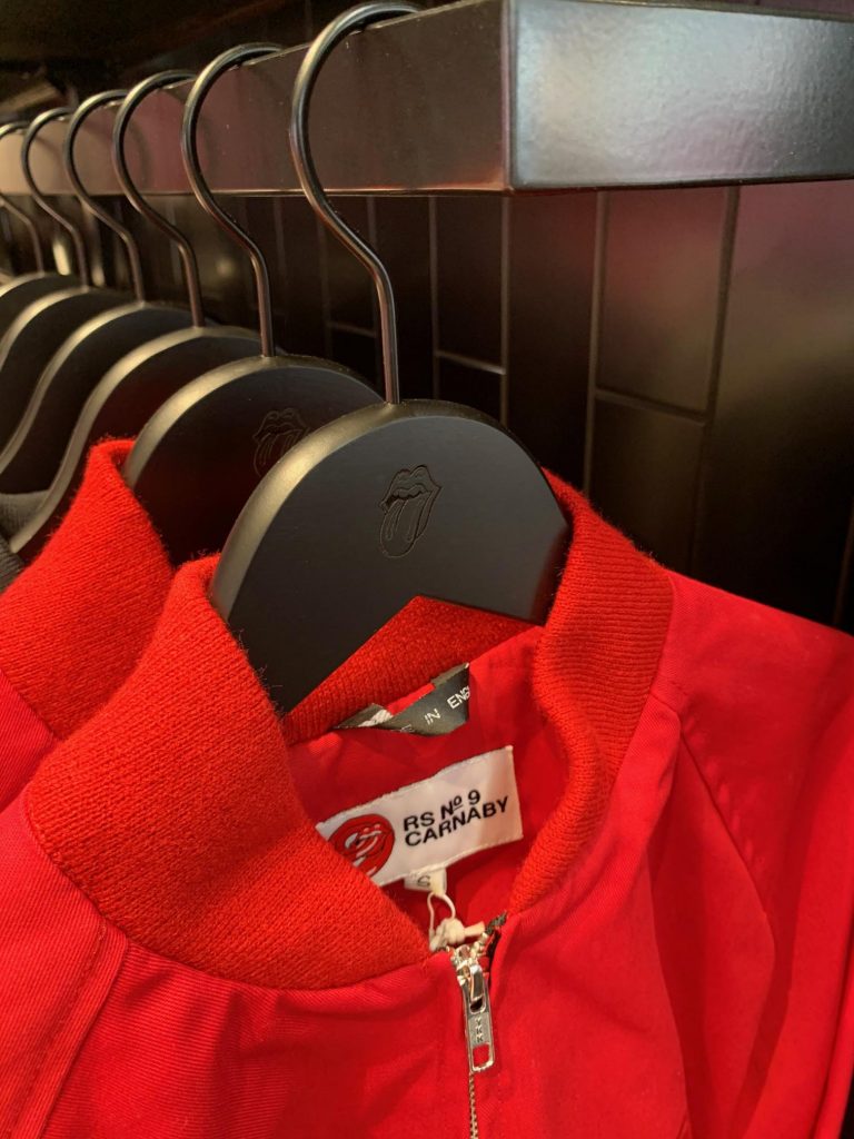 HOL Group A red jacket hangs on a branded hanger featuring the Rolling Stones' iconic tongue and lips logo. The jacket tag reads "RS No 9 Carnaby," indicating its association with the legendary rock band’s fashion line. Other jackets are visible in the background, echoing the music legacy of the Rolling Stones.