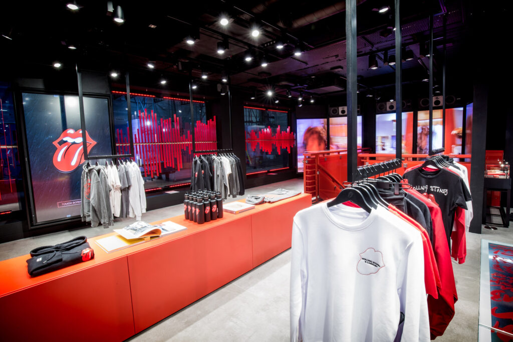 HOL Group A modern retail store celebrating the rock legends, Rolling Stones. The space features red shelving and hanging racks displaying various clothing items, including shirts with the iconic tongue and lips logo. Bright, colorful lighting and a screen displaying soundwave patterns decorate the space.