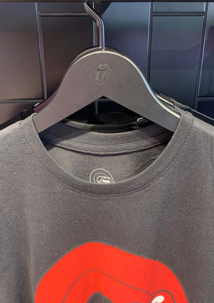 HOL Group A black t-shirt with a red and white graphic of a mouth and tongue, reminiscent of the iconic Rolling Stones logo, is displayed on a black hanger. The hanger features a similar tongue logo embossed at the center. The background consists of black panels, adding to the rock band aesthetic.