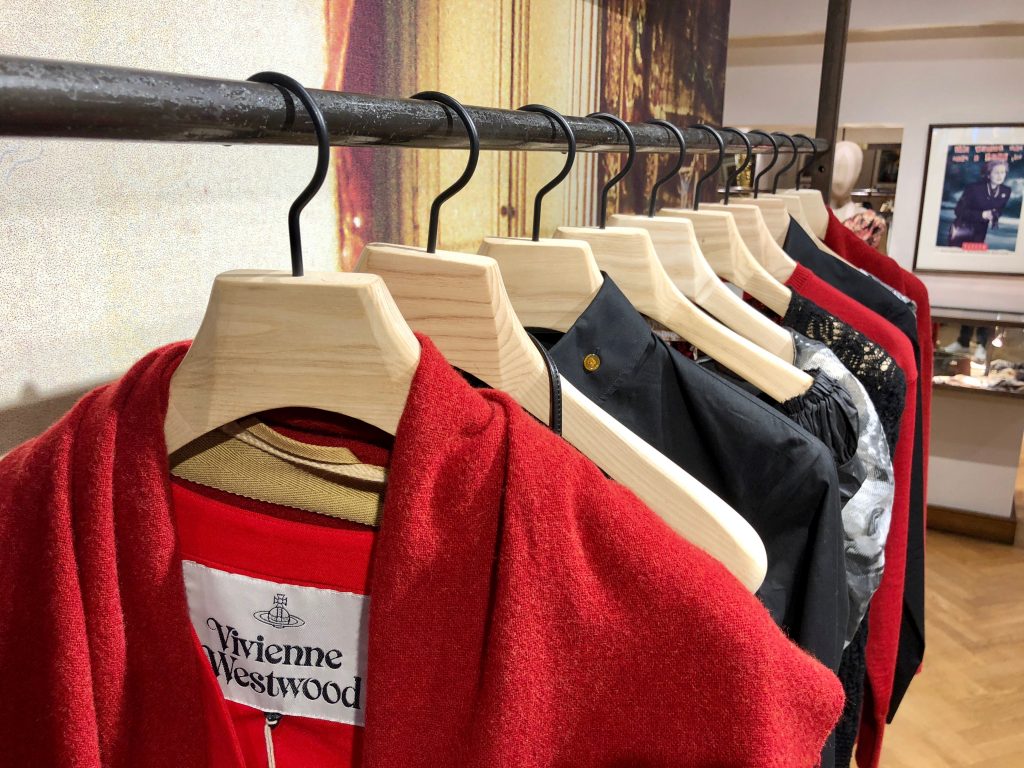 HOL Group A clothing rack displays several items of clothing, primarily featuring red and black garments. A tag on the first item, a striking red piece, proudly reads "Vivienne Westwood." The background includes a framed picture and a glimpse of a stylish shop interior.