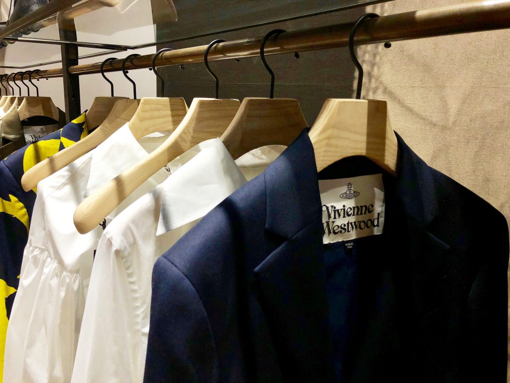 HOL Group A clothing rack holds multiple garments on wooden hangers, including a navy blazer by Vivienne Westwood and various white and patterned shirts. The background displays part of a fabric wall and a dimly lit space.